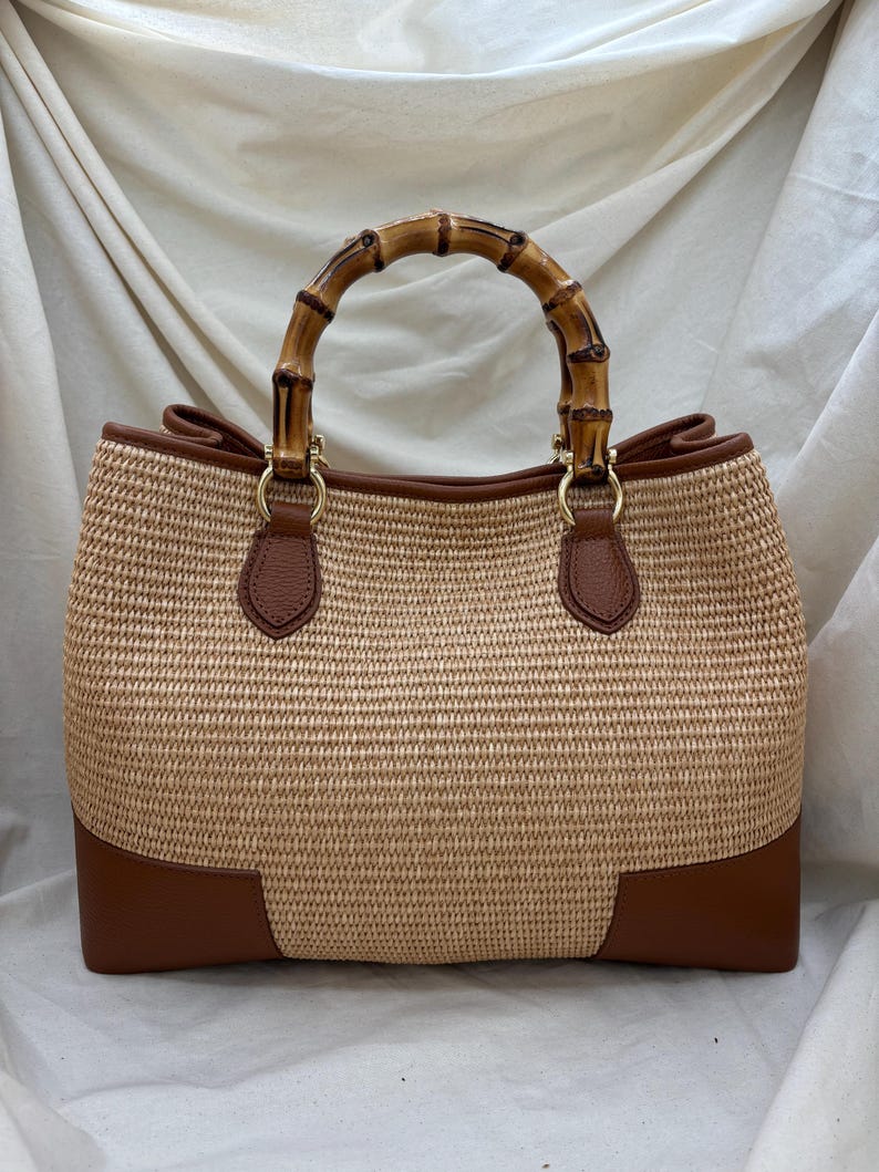 Pode incluir: Uma bolsa tote tecida com uma al&ccedil;a de bambu. A bolsa apresenta um corpo tecido castanho claro com detalhes em couro castanho nos cantos inferiores e nas fixa&ccedil;&otilde;es da al&ccedil;a. A al&ccedil;a &eacute; feita de bambu curvo.