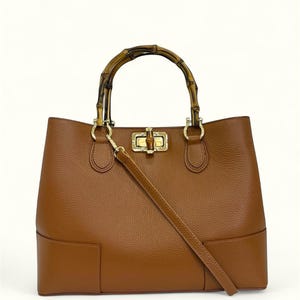 May include: A brown leather handbag with a textured surface, featuring a curved bamboo handle and a gold-toned metal turn-lock closure. The bag includes a detachable, matching brown leather shoulder strap attached to gold-toned hardware rings.