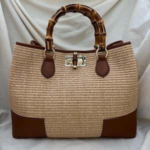 Pode incluir: Uma mala tote de r&aacute;fia tecida com uma al&ccedil;a de bambu. A mala apresenta detalhes em couro castanho na parte inferior e na borda, e um fecho dourado. A al&ccedil;a &eacute; feita de bambu curvo, adicionando um toque natural ao design.