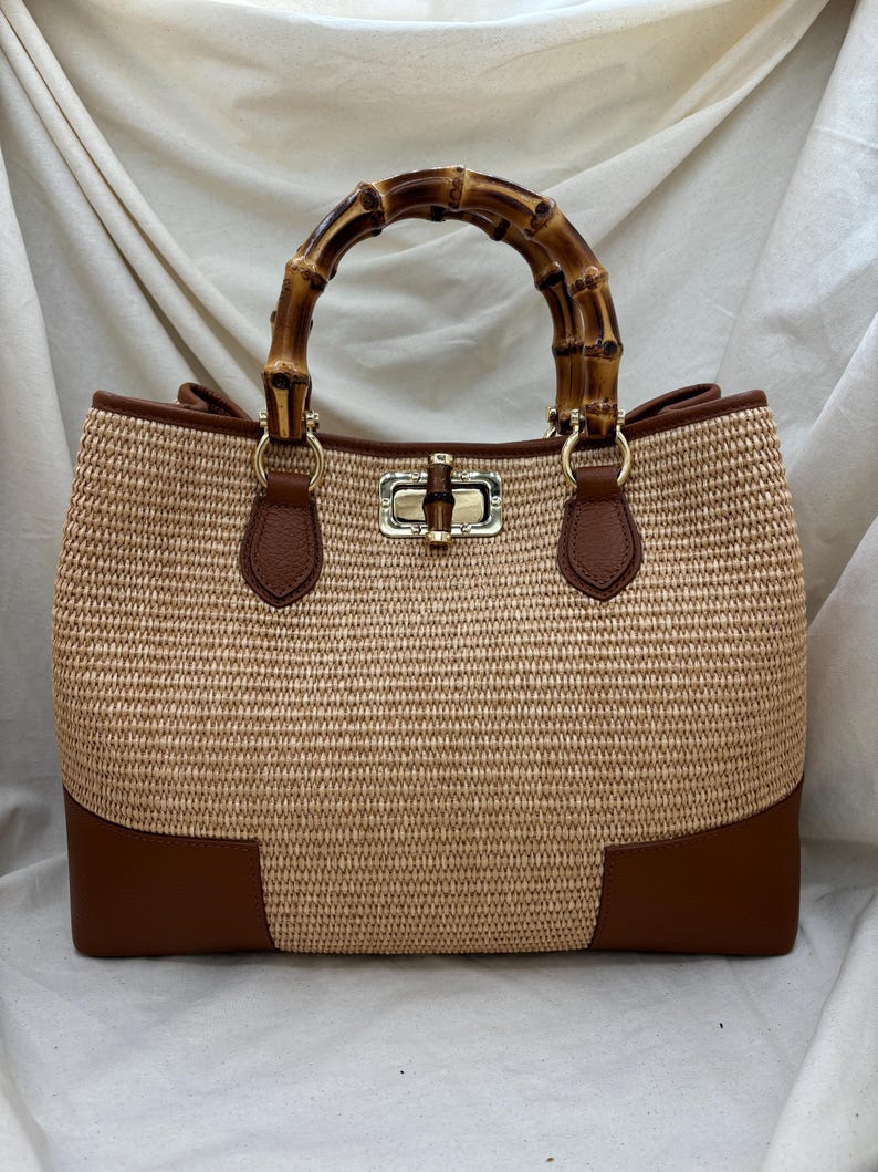 Pode incluir: Uma mala tote de r&aacute;fia tecida com uma al&ccedil;a de bambu. A mala apresenta detalhes em couro castanho na parte inferior e na borda, e um fecho dourado. A al&ccedil;a &eacute; feita de bambu curvo, adicionando um toque natural ao design.