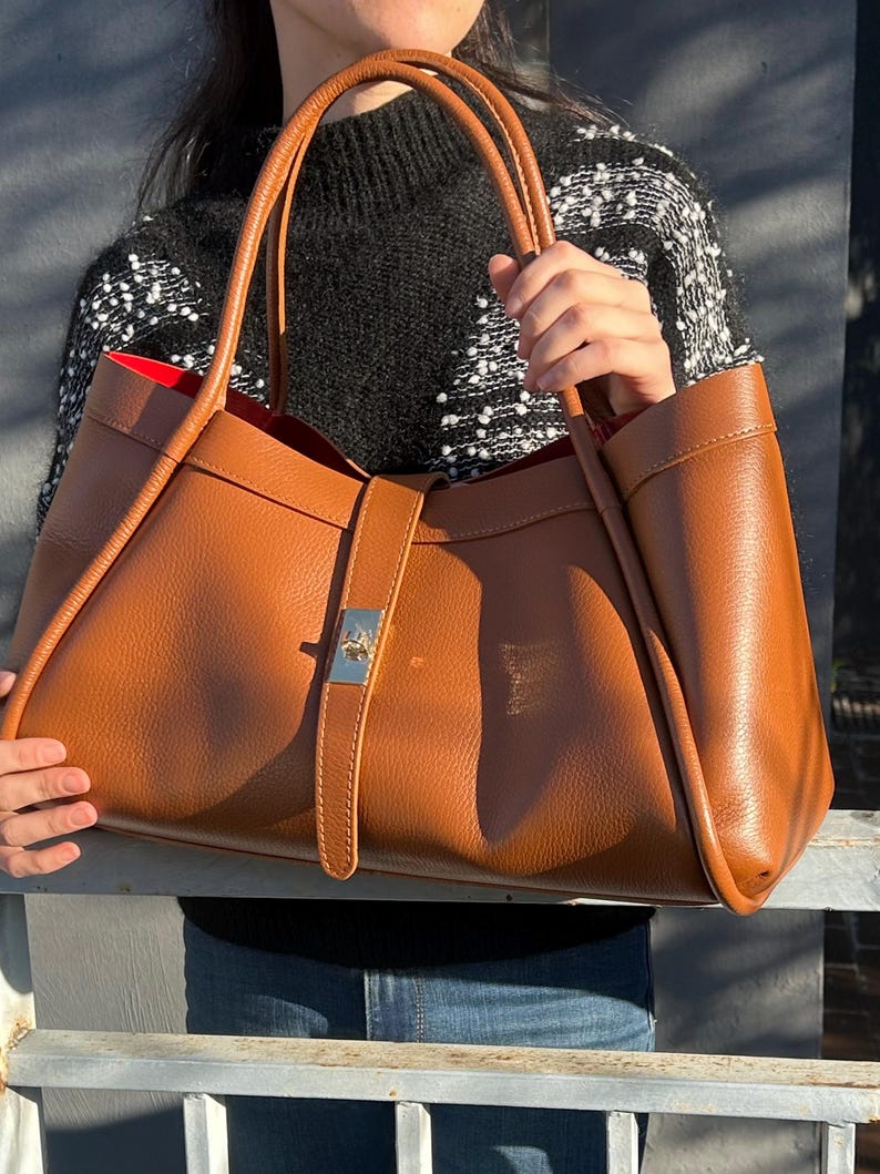 K&ouml;nnte beinhalten: Eine gro&szlig;e, braune Ledertasche mit rotem Innenfutter. Die Tasche hat zwei Griffe und einen silberfarbenen Schnallenverschluss. Die Tasche wird von einer Person gehalten.