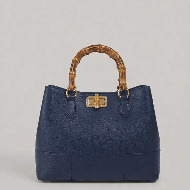 May include: A navy blue handbag with a textured surface, featuring a curved bamboo handle and a gold-toned metal turn-lock closure. The bag has a structured base and is attached to the handle with gold-toned metal hardware rings.