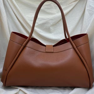 K&ouml;nnte beinhalten: Eine braune Ledertasche mit rotem Innenfutter. Die Tasche hat zwei lange Henkel und eine rechteckige Form mit einem Verschluss. Die Tasche ist aus glattem, strukturiertem Leder gefertigt.