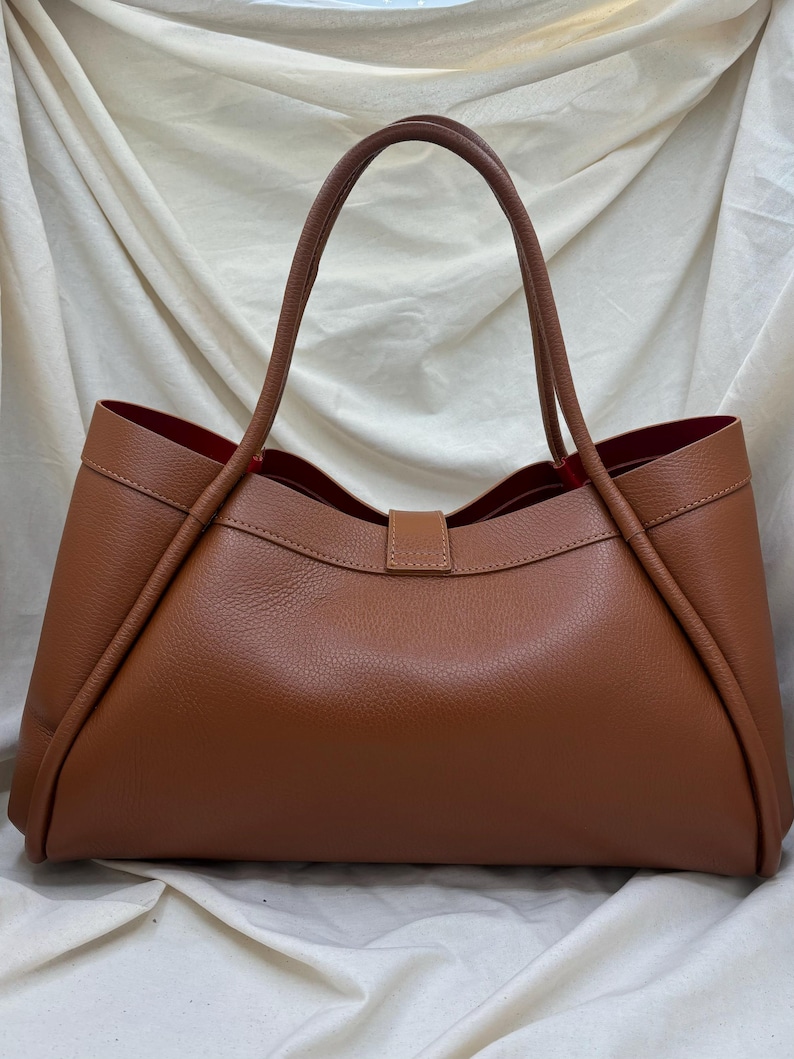 K&ouml;nnte beinhalten: Eine braune Ledertasche mit rotem Innenfutter. Die Tasche hat zwei lange Henkel und eine rechteckige Form mit einem Verschluss. Die Tasche ist aus glattem, strukturiertem Leder gefertigt.
