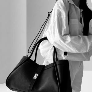 K&ouml;nnte beinhalten: Eine schwarze Ledertasche mit silbernem Verschluss und abnehmbarem Schulterriemen. Die Tasche hat zwei obere Griffe und ein strukturiertes Design. Das Bild ist in Schwarzwei&szlig;.