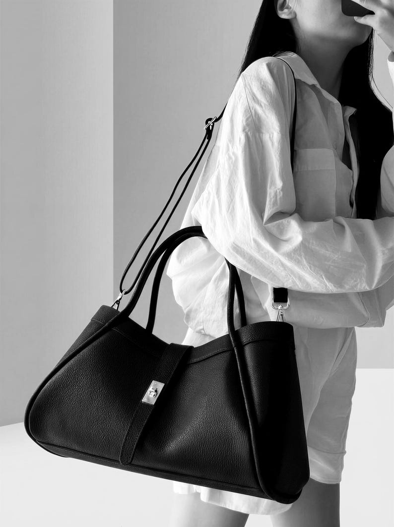 K&ouml;nnte beinhalten: Eine schwarze Ledertasche mit silbernem Verschluss und abnehmbarem Schulterriemen. Die Tasche hat zwei obere Griffe und ein strukturiertes Design. Das Bild ist in Schwarzwei&szlig;.