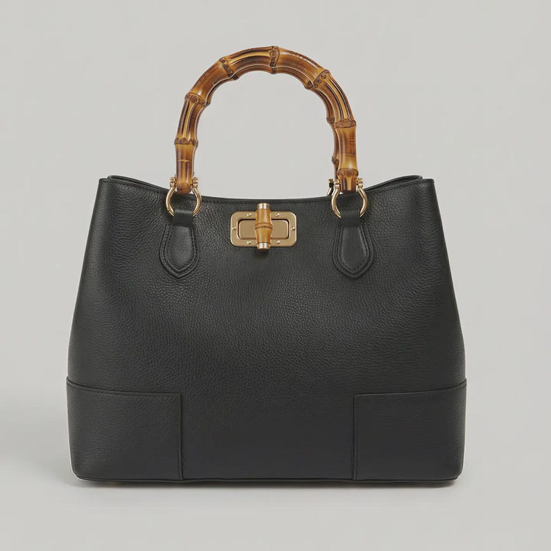 May include: A black leather handbag featuring a textured surface, a curved bamboo handle, and a gold-toned metal turn-lock closure. The bag has a structured base and is attached to the handle with gold-toned metal rings and leather tabs.