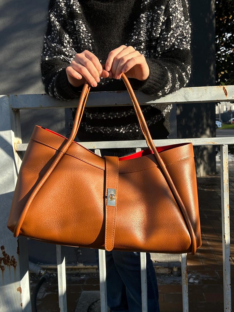 K&ouml;nnte beinhalten: Eine gro&szlig;e, braune Ledertasche mit rotem Innenfutter. Die Tasche hat lange Henkel und einen Riemenverschluss mit silberfarbener Schnalle. Die Tasche wird von einer Person gehalten, die einen schwarz-wei&szlig; gemusterten Pullover tr&auml;gt.