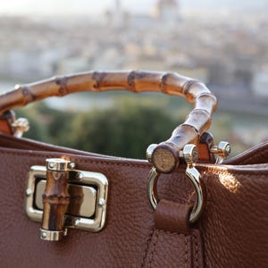 May include: A brown leather handbag featuring a curved bamboo handle and a rectangular bamboo turn-lock closure. The bag is accented with polished silver-tone metal hardware, including circular rings that connect the handle to the textured leather body.