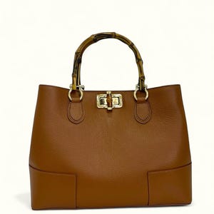 May include: A tan leather handbag with a textured surface, featuring a rounded bamboo handle attached with gold-toned metal hardware. The bag includes a central gold-toned metal turn-lock closure mechanism on the front panel and a structured base.
