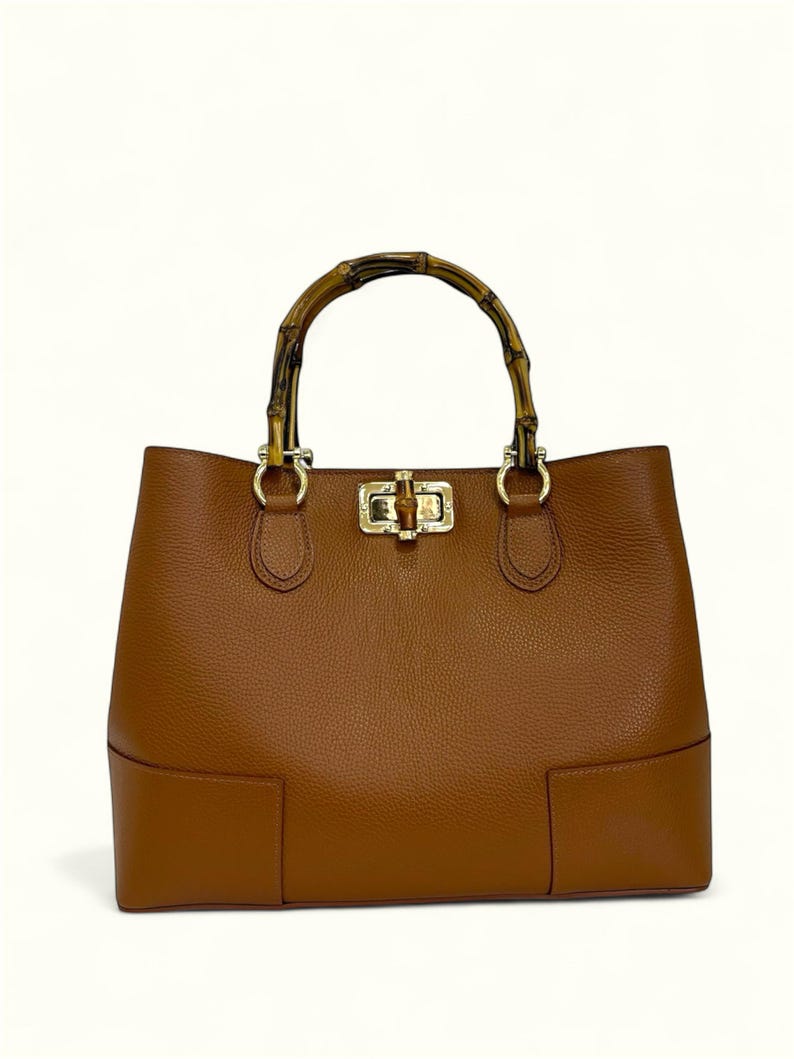 May include: A tan leather handbag with a textured surface, featuring a rounded bamboo handle attached with gold-toned metal hardware. The bag includes a central gold-toned metal turn-lock closure mechanism on the front panel and a structured base.