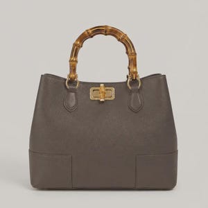 May include: A structured grey handbag with a textured surface, featuring a distinctive arched bamboo handle and a central gold-toned metal turn-lock closure. The bag has a wide base and is attached to the handle with gold-toned metal hardware.