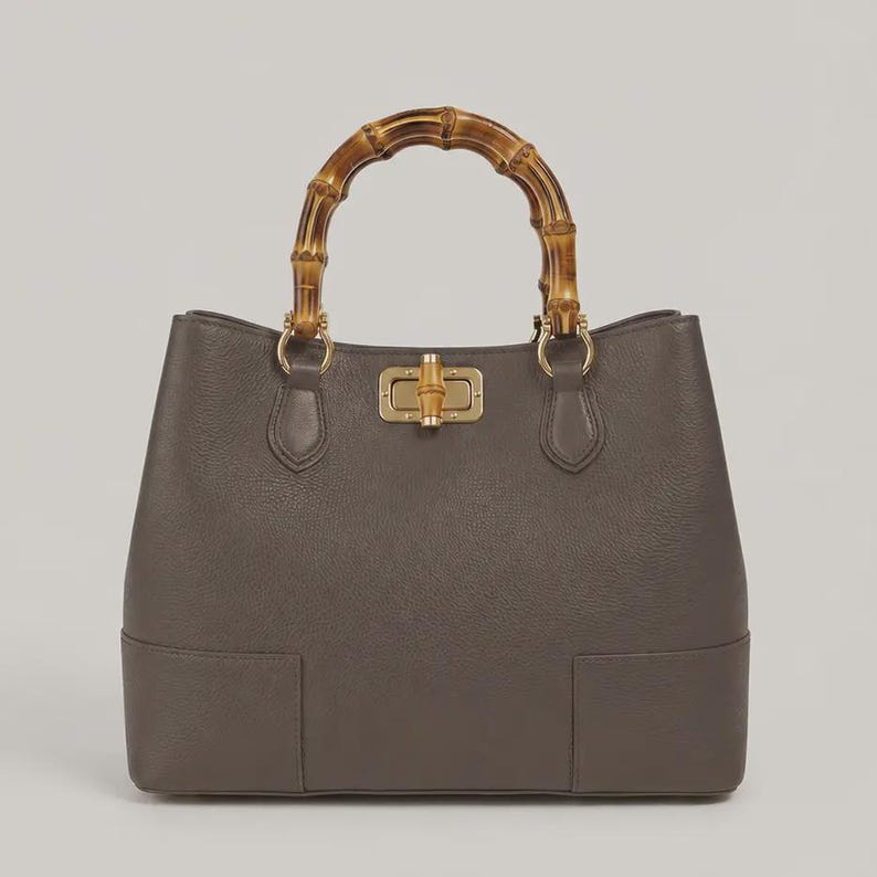 May include: A structured grey handbag with a textured surface, featuring a distinctive arched bamboo handle and a central gold-toned metal turn-lock closure. The bag has a wide base and is attached to the handle with gold-toned metal hardware.