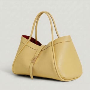 K&ouml;nnte beinhalten: Eine hellgelbe Ledertasche mit rotem Innenfutter. Die Tasche hat zwei lange Henkel und einen Riemen mit einem goldfarbenen Verschluss. Das Design der Tasche ist strukturiert mit abgerundeten Kanten.