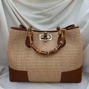 Pode incluir: Uma bolsa tote bege tecida com detalhes em couro castanho e uma al&ccedil;a de bambu. A bolsa tem formato retangular com fecho dourado e cantos em couro castanho. A al&ccedil;a &eacute; feita de bambu curvo.