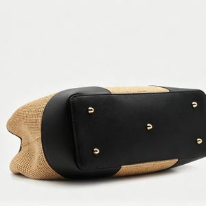 Pode incluir: Uma bolsa tote bege tecida com detalhes em couro preto. A bolsa apresenta uma base retangular com p&eacute;s de metal dourado. A parte superior da bolsa tamb&eacute;m &eacute; de couro preto, e as laterais s&atilde;o de material bege tecido.