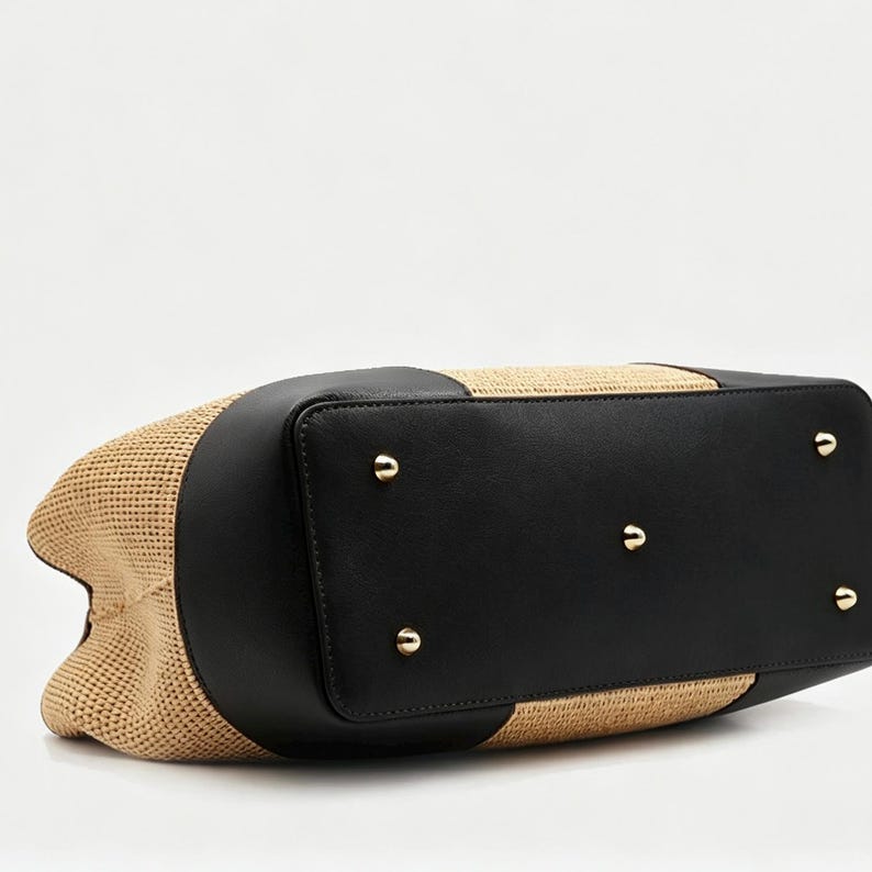 Pode incluir: Uma bolsa tote bege tecida com detalhes em couro preto. A bolsa apresenta uma base retangular com p&eacute;s de metal dourado. A parte superior da bolsa tamb&eacute;m &eacute; de couro preto, e as laterais s&atilde;o de material bege tecido.