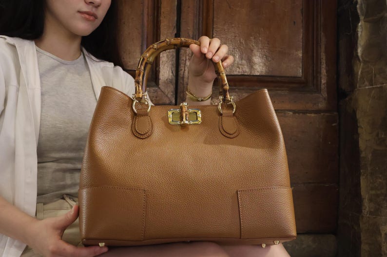 May include: A tan leather handbag with a textured surface, featuring a curved bamboo handle and a gold-toned metal turn-lock closure. The bag has a structured rectangular base with visible stitching details and metallic feet on the bottom corners.