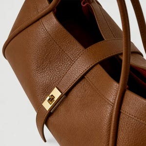 K&ouml;nnte beinhalten: Nahaufnahme einer braunen Lederhandtasche mit goldenem Verschluss. Die Tasche hat eine strukturierte Oberfl&auml;che und ein rotes Innenfutter. Der Riemen ist um die Tasche gewickelt und mit einem goldenen Verschluss befestigt.
