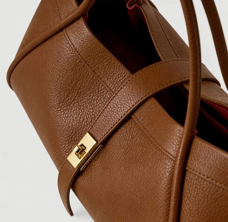 K&ouml;nnte beinhalten: Nahaufnahme einer braunen Lederhandtasche mit goldenem Verschluss. Die Tasche hat eine strukturierte Oberfl&auml;che und ein rotes Innenfutter. Der Riemen ist um die Tasche gewickelt und mit einem goldenen Verschluss befestigt.