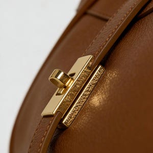 K&ouml;nnte beinhalten: Nahaufnahme einer braunen Ledertasche mit einem goldfarbenen Verschluss. Der Verschluss ist rechteckig mit einem gebogenen Hebel und einem strukturierten Dekorationselement. Der Lederriemen ist gen&auml;ht und hat einen passenden Braunton.