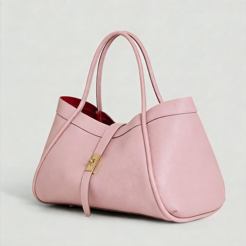 K&ouml;nnte beinhalten: Eine hellrosa Ledertasche mit strukturiertem Design. Die Tasche hat zwei lange Griffe, einen goldfarbenen Verschluss und ein rotes Innenfutter. Die Tasche hat einen Riemen, der mit einem goldfarbenen Verschluss befestigt wird.