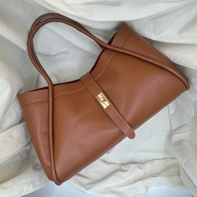 K&ouml;nnte beinhalten: Eine braune Ledertasche mit rechteckiger Form und einem goldfarbenen Verschluss. Die Tasche hat zwei lange Griffe und einen Riemen, der die Vorderseite sichert. Der Innenraum ist in einem dunkleren Rotton gehalten.