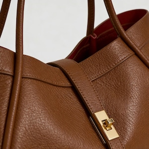 K&ouml;nnte beinhalten: Nahaufnahme einer braunen Ledertasche mit goldenem Verschluss. Die Tasche hat eine strukturierte Oberfl&auml;che und ein rotes Innenfutter. Die Griffe sind oben sichtbar, und ein Riemen mit Schnalle sichert die Tasche.