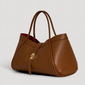 K&ouml;nnte beinhalten: Braune Ledertasche mit rotem Innenraum. Die Tasche hat zwei lange Henkel und einen Riemen mit goldfarbenem Verschluss. Das Design der Tasche ist trapezf&ouml;rmig, mit einer strukturierten Lederoberfl&auml;che.
