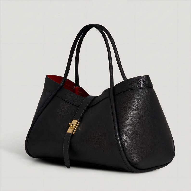 K&ouml;nnte beinhalten: Eine schwarze Ledertasche mit rotem Innenfutter. Die Tasche hat zwei schwarze Griffe und einen goldfarbenen Verschluss. Das Design ist strukturiert und elegant, geeignet f&uuml;r den t&auml;glichen Gebrauch.
