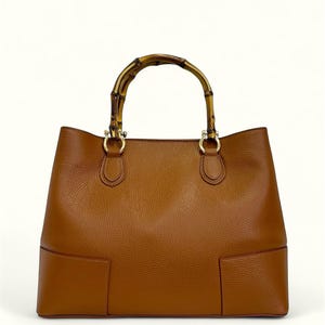May include: A tan leather handbag with a textured surface and a rounded bamboo handle. The bag features gold-toned metal hardware connecting the handle to the body, with a structured base and clean, minimalist lines throughout the design.