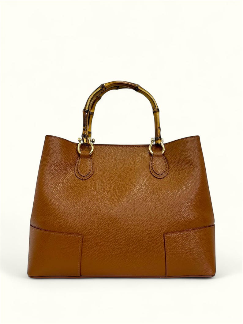 May include: A tan leather handbag with a textured surface and a rounded bamboo handle. The bag features gold-toned metal hardware connecting the handle to the body, with a structured base and clean, minimalist lines throughout the design.