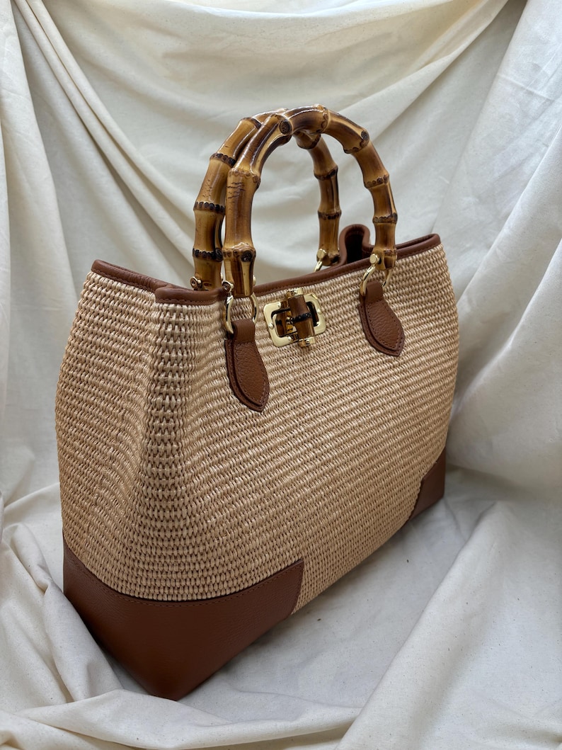 Pode incluir: Uma mala tote de palha tecida com uma al&ccedil;a de bambu. A mala apresenta detalhes em couro castanho nos cantos e na borda, e um fecho dourado. A mala &eacute; de cor bege claro.