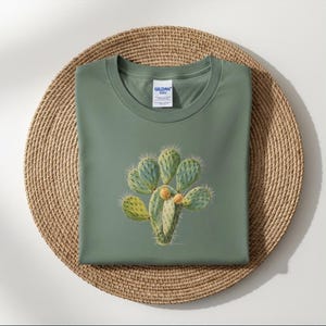 Può includere: Una maglietta verde salvia con un'illustrazione dettagliata di un fico d'India. Il cactus ha pale verdi e verde-bluastre con frutti giallo-arancio. La maglietta è piegata su un sottobicchiere rotondo, intrecciato e marrone.