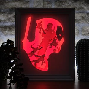 Può includere: Una scatola luminosa rettangolare incorniciata che presenta una silhouette retroilluminata in rosso vibrante del personaggio Deadpool. Il design incorpora diverse sagome nere più piccole del personaggio in varie pose d'azione, tra cui impugnare spade e armi.