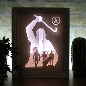 May include: A framed, illuminated artwork showing a silhouette of a person with a crowbar, and the Lambda symbol. The scene depicts figures walking in a city, with the text "CITY 17". The frame is light-coloured.