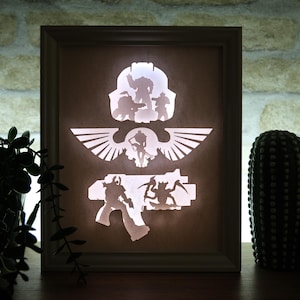May include: A framed, illuminated artwork featuring cut-out silhouettes of figures and symbols. The design includes a helmet, winged emblem, and weapon, all in white against a warm, brown background. The frame is a light brown color.