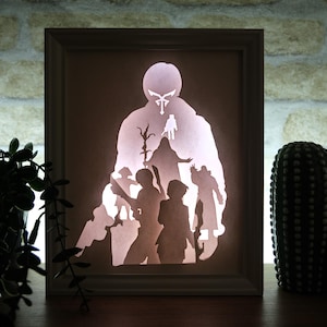 May include: A framed, backlit paper cut art piece showing a central figure with other figures around it. The artwork is in a light-coloured wooden frame, with a brick wall background. Decorative plants and cacti are in the foreground.