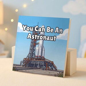 May include: A square book cover featuring a cartoon illustration of a white rocket on a launch pad against a blue sky with clouds. The text You Can Be An Astronaut is printed in bold black letters with a white outline across the center of the image.