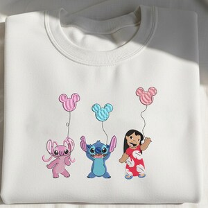 K&ouml;nnte beinhalten: Ein wei&szlig;es Sweatshirt mit Rundhalsausschnitt und einer Stickerei, die einen rosa Au&szlig;erirdischen, einen blauen Au&szlig;erirdischen und ein M&auml;dchen in einem roten Kleid zeigt. Jede Figur h&auml;lt eine Schnur mit einem Mickey-Maus-Ballon in passender Farbe.