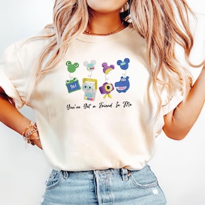 May include: A cream-colored t-shirt featuring a graphic print of four cartoon characters with spring-like attachments and the text "You've Got a Friend In Me" in black script. The characters include a green frog, a white robot, a purple camera, and a blue hippo.