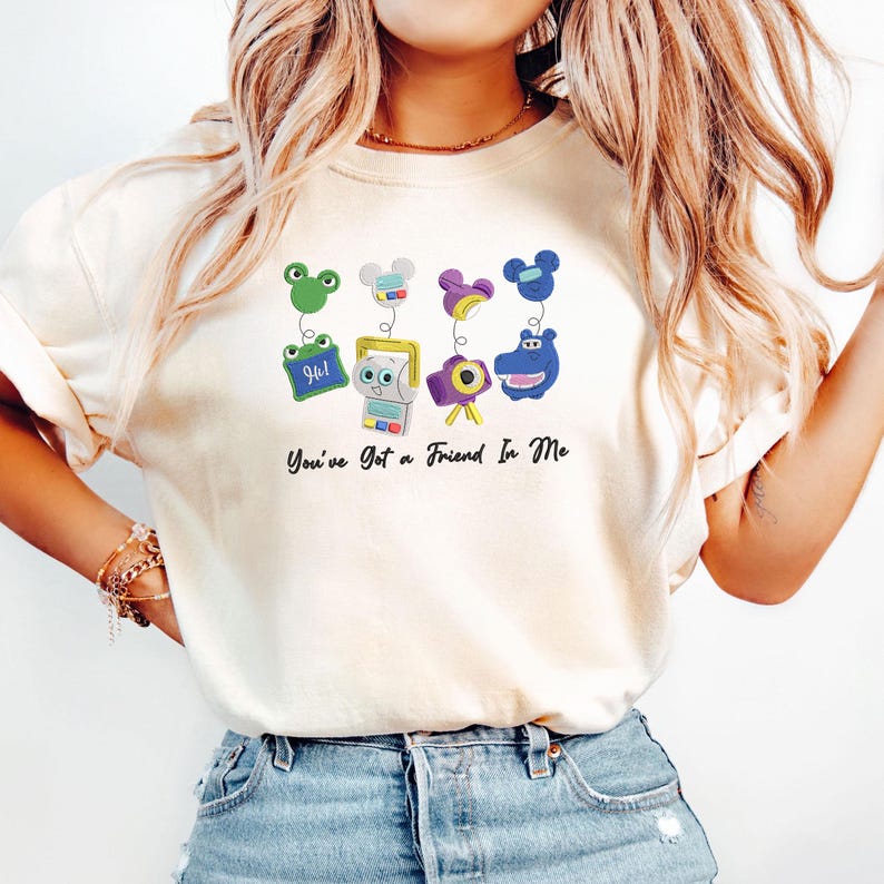 May include: A cream-colored t-shirt featuring a graphic print of four cartoon characters with spring-like attachments and the text "You've Got a Friend In Me" in black script. The characters include a green frog, a white robot, a purple camera, and a blue hippo.