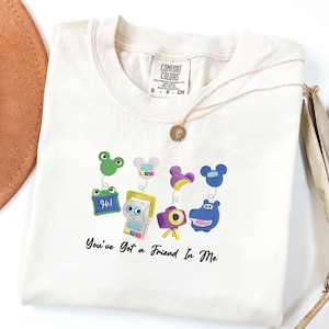 May include: A cream-colored short-sleeve t-shirt featuring a graphic of four colorful characters on springs, including a green frog, a gray device, a purple camera, and a blue hippo. The text "You've Got a Friend In Me" is printed in black below.