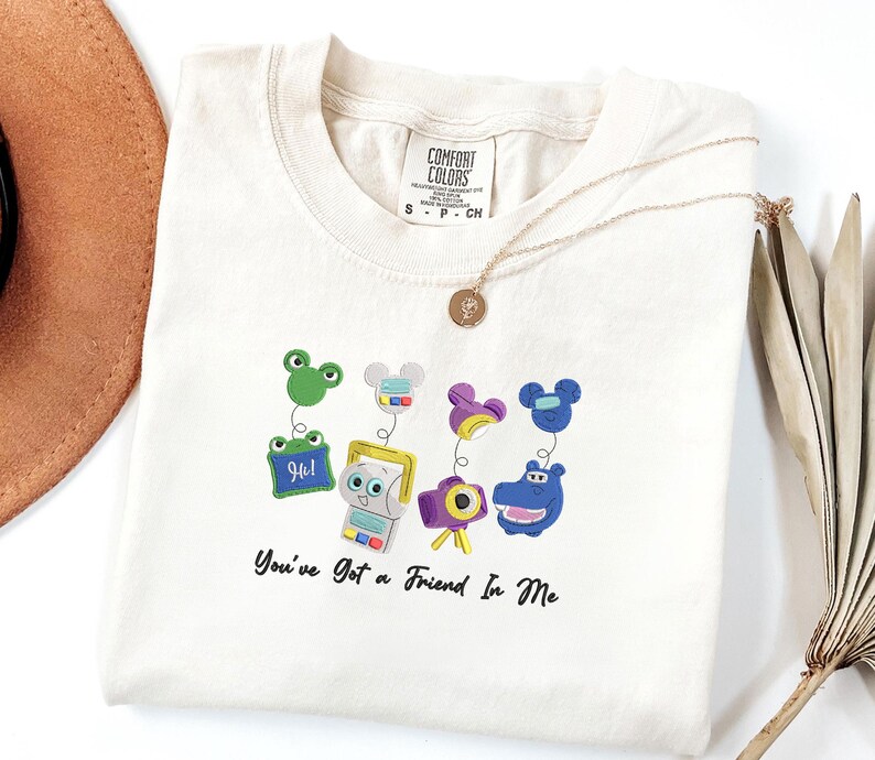 May include: A cream-colored short-sleeve t-shirt featuring a graphic of four colorful characters on springs, including a green frog, a gray device, a purple camera, and a blue hippo. The text "You've Got a Friend In Me" is printed in black below.