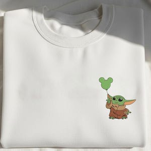 May include: A white crewneck sweatshirt featuring a small, embroidered graphic on the right side. The design depicts a green, large-eared character wearing a brown robe, holding a green balloon shaped like a three-circle mouse head silhouette.