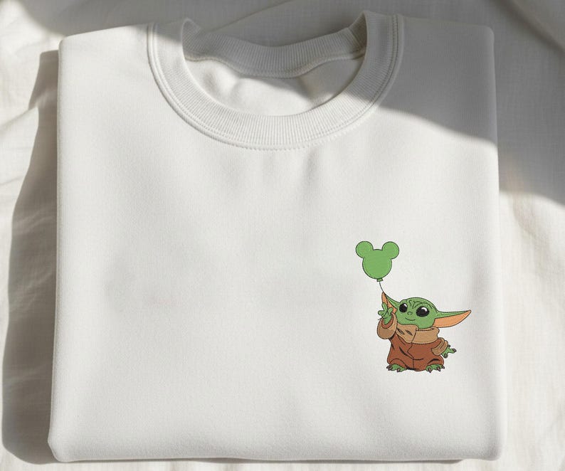 May include: A white crewneck sweatshirt featuring a small, embroidered graphic on the right side. The design depicts a green, large-eared character wearing a brown robe, holding a green balloon shaped like a three-circle mouse head silhouette.
