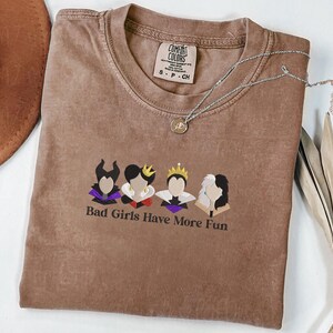May include: A brown Comfort Colors t-shirt with the phrase "Bad Girls Have More Fun" and a graphic of four cartoon characters. A silver necklace with a small pendant is on the shirt.