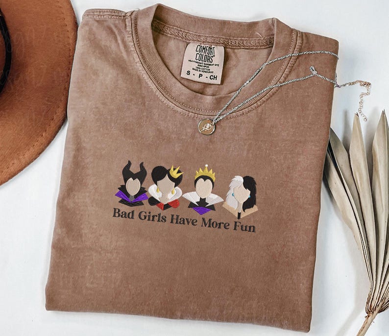 May include: A brown Comfort Colors t-shirt with the phrase "Bad Girls Have More Fun" and a graphic of four cartoon characters. A silver necklace with a small pendant is on the shirt.