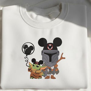 Op de afbeelding: Een wit sweatshirt met ronde hals en een grafische print van een klein groen wezen in een bruine mantel en een figuur in een grijs harnas, beide met zwarte Mickey Mouse-oren. Het kleine wezen houdt een zwarte Mickey Mouse-ballon vast.