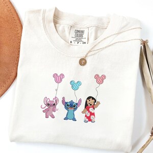 K&ouml;nnte beinhalten: Ein cremefarbenes Kurzarm-T-Shirt mit einer Stickerei von zwei blauen und rosa au&szlig;erirdischen Charakteren und einem M&auml;dchen, das drei pinke und blaue Luftballons in Mickey-Mouse-Form h&auml;lt. Das Shirt hat ein Comfort Colors Markenetikett am Hals.
